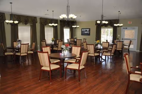 A dining room with round tables and upholstered chairs in a senior living facility.