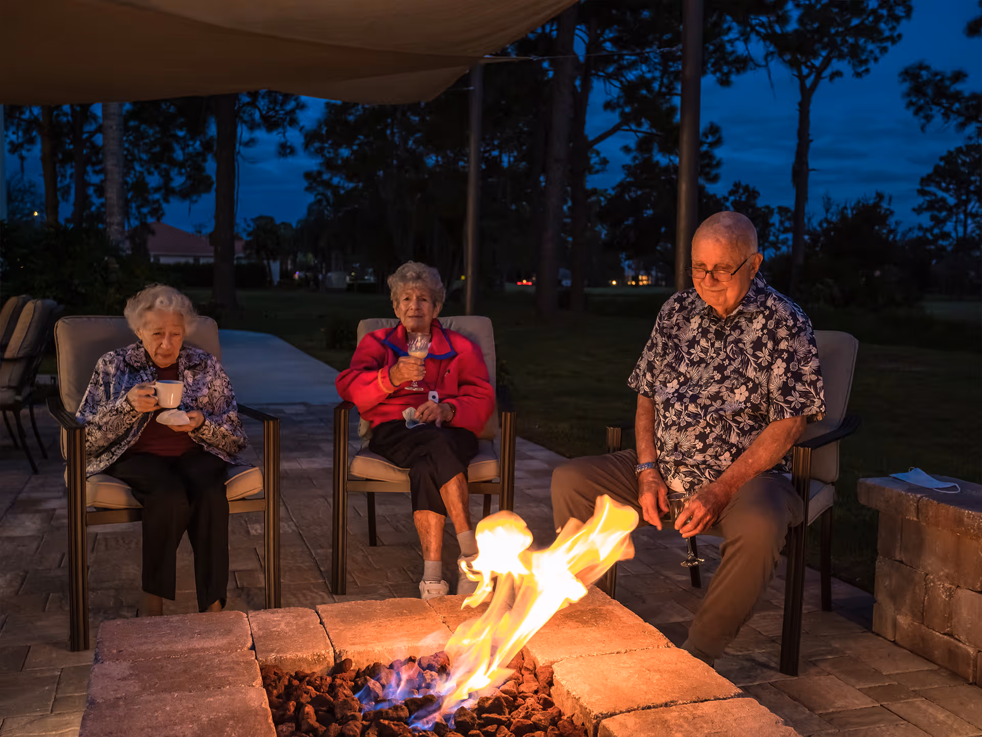 Three elderly people sitting around an outdoor fire pit at dusk. Two women and one man are seated on cushioned chairs on a paved patio. One woman is holding a cup and saucer, the other woman is holding a glass, and the man is also holding a glass. Trees and a darkening sky are visible in the background.