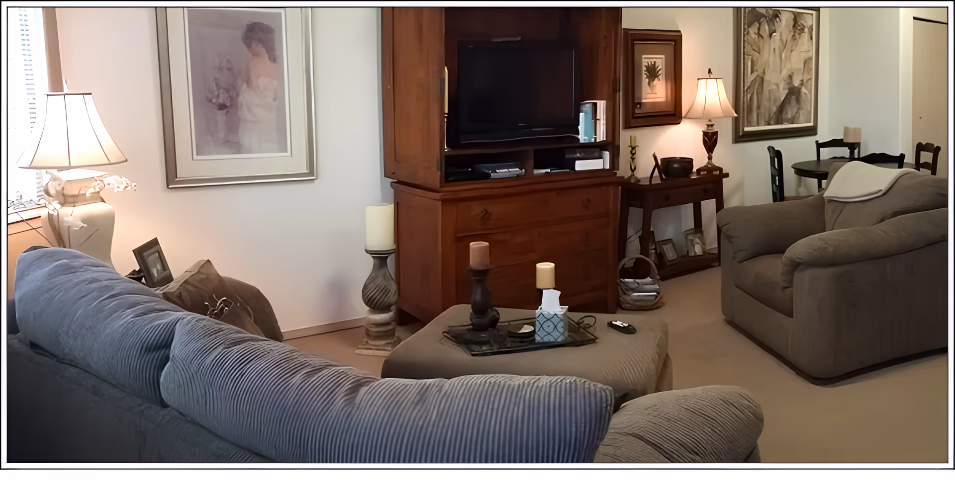 A cozy living room with a large gray sofa and matching armchair. A wooden entertainment center holds a flat-screen TV. There are decorative candles on a cushioned ottoman in the center. The room is softly lit by table lamps and decorated with framed artwork on the walls. In the background, a small dining table with chairs is visible.