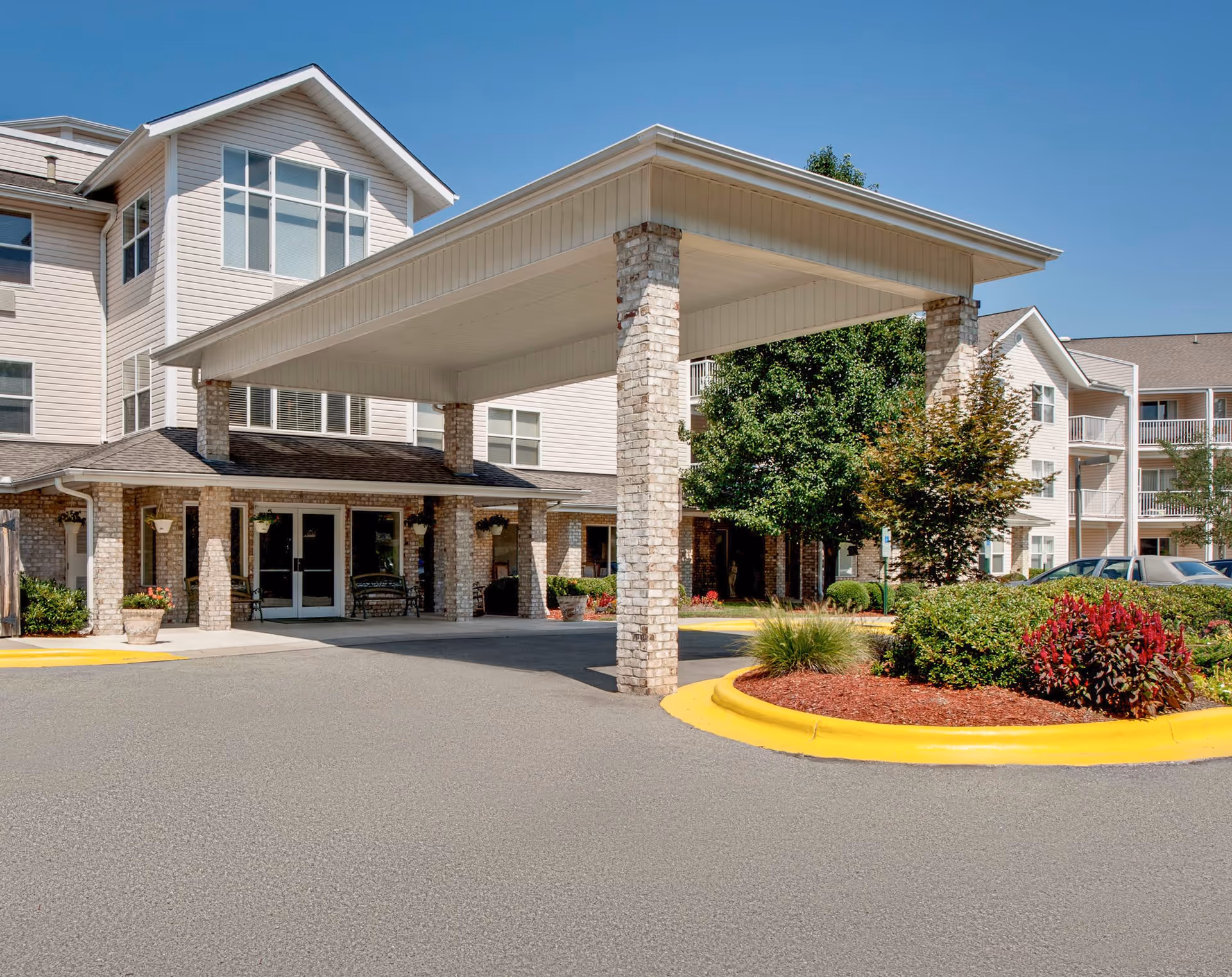 Exterior view of Solista Durham by Cogir senior living facility showing the main entrance with a covered drop-off area supported by brick columns, landscaped greenery, and a clear blue sky.