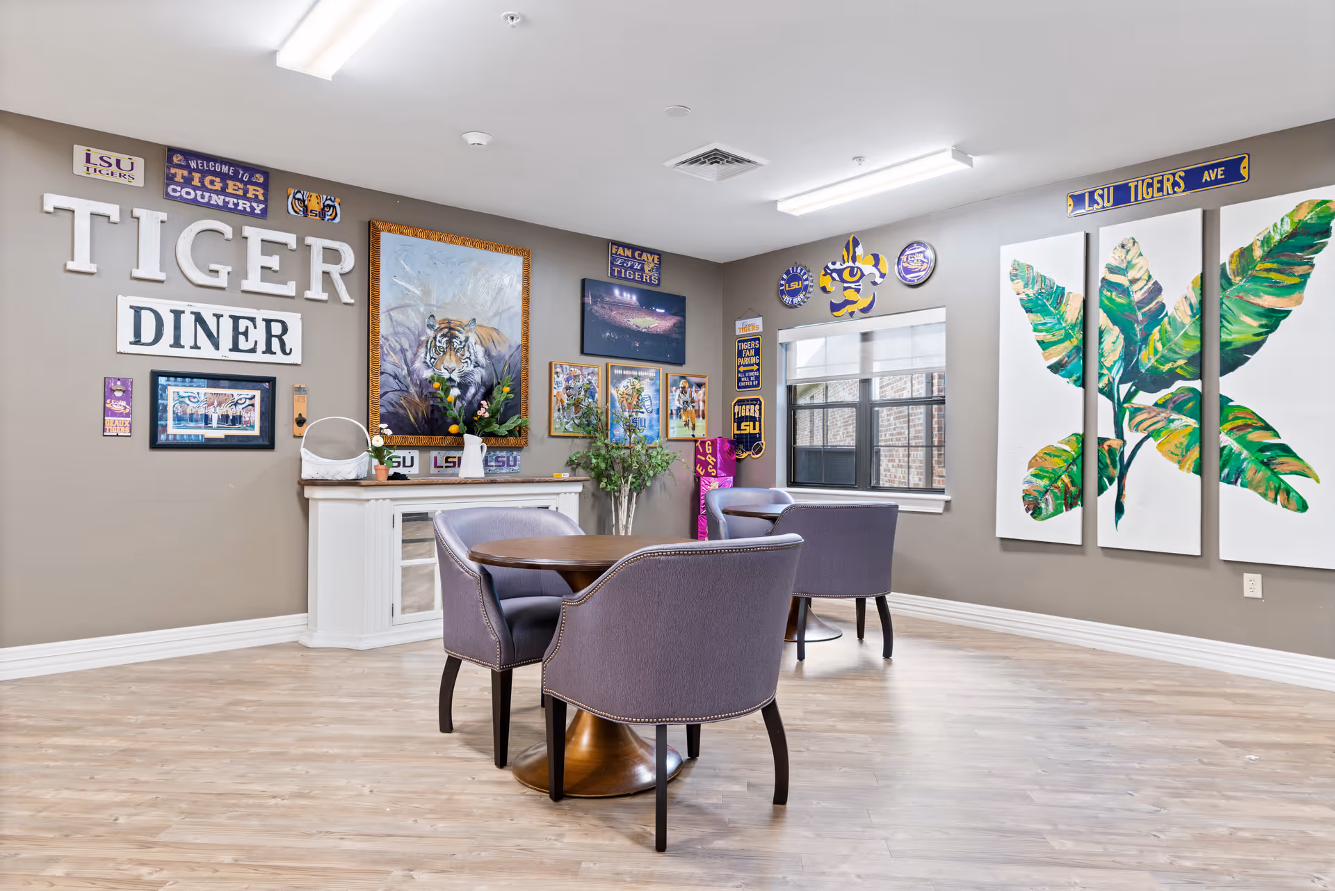 Interior view of a dining area named 'Tiger Diner' decorated with LSU Tigers memorabilia including signs, paintings, and banners. The room has a wooden floor, a round wooden table with four purple upholstered chairs, a white cabinet with plants and flowers, and a window with white blinds. The walls are painted beige and adorned with various LSU-themed decorations and a large three-panel painting of green leaves.