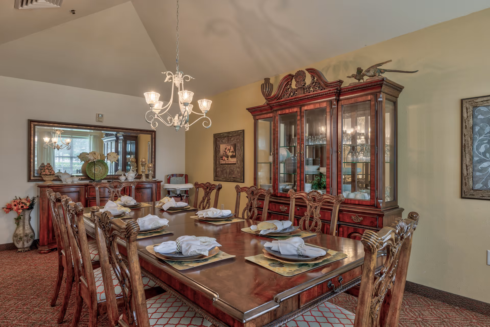 A formal dining room with a large wooden dining table set for eight people with white napkins and plates. The room features ornate wooden chairs, a matching wooden china cabinet filled with glassware, a sideboard with decorative items, a large mirror on the wall, a chandelier hanging from the ceiling, and framed artwork on the walls.