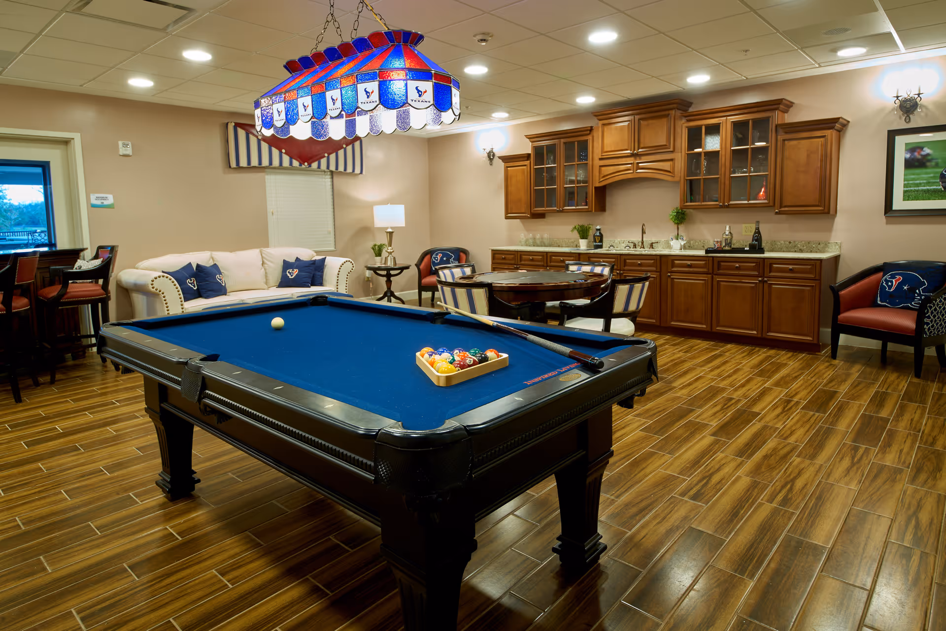 A recreational room with a blue felt pool table in the center, a white couch with blue pillows, a round table with chairs, wooden cabinets with a countertop and sink, and a stained glass hanging light fixture above the pool table. The room has wood-look tile flooring and beige walls.