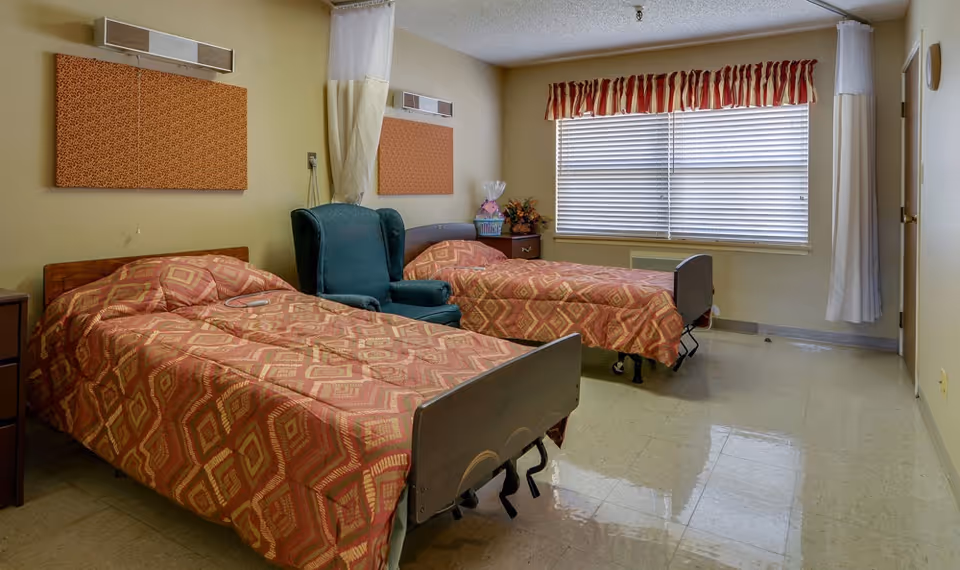 A simple bedroom with two single beds covered in patterned bedspreads, a green upholstered armchair between the beds, a window with blinds and a red and white valance, and beige walls with two rectangular wall-mounted panels above the beds.