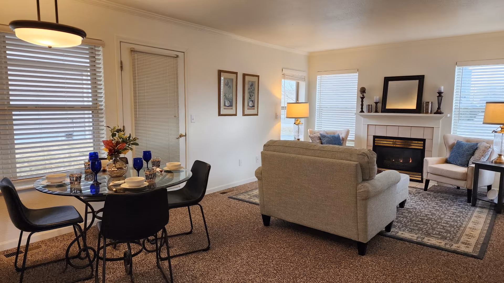 A cozy living and dining area with a round glass dining table set with blue glasses and white dishes, surrounded by four black chairs. The living room features a beige sofa facing a white fireplace with a black-framed mirror and decorative candles on the mantel. Two beige armchairs with blue cushions flank the fireplace, and the room is lit by table lamps and natural light from multiple windows with blinds.