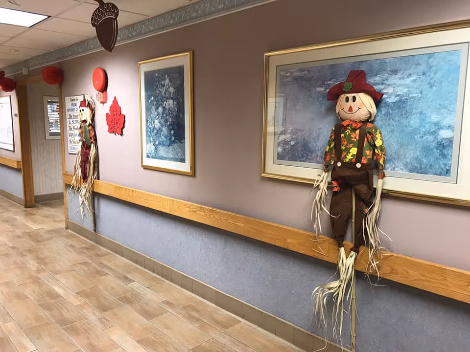 Hallway in a senior living facility decorated with autumn scarecrows, framed artwork, and wooden handrails.