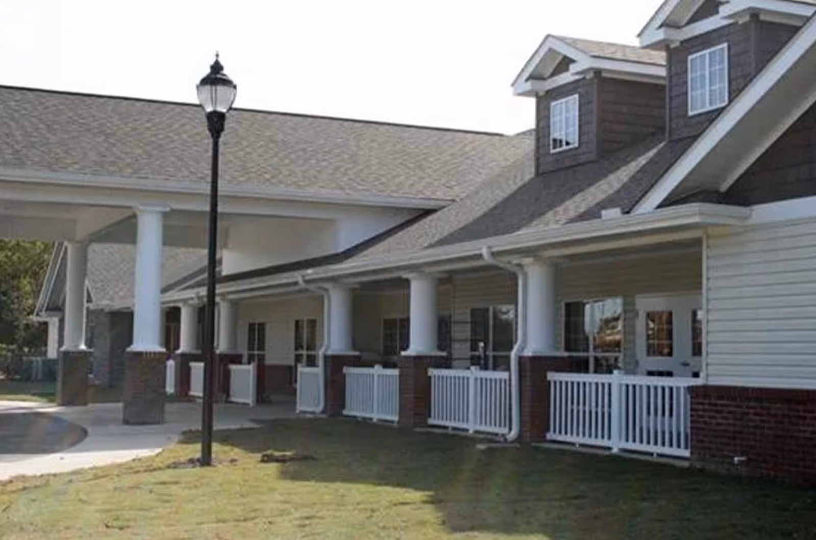 Exterior view of a single-story building with a covered porch supported by white columns and white railings. The building has a gray shingled roof with dormer windows and a brick base. A black streetlamp is visible on the lawn in front of the building.