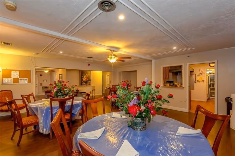 Dining room with round tables covered in blue plastic tablecloths, floral centerpieces, and wooden chairs with a pass-through to the kitchen.