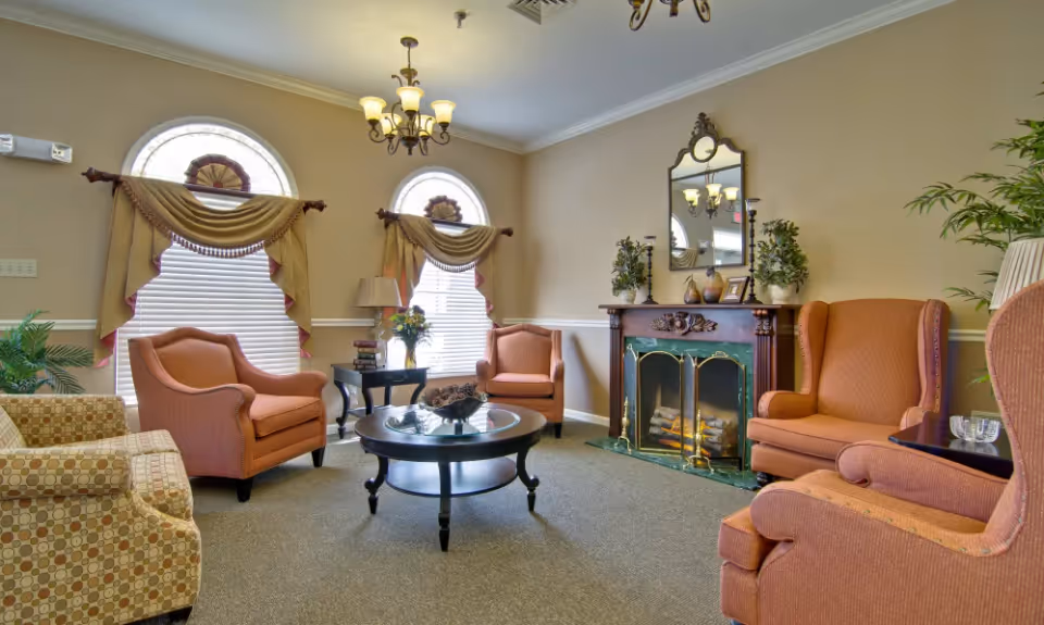 A cozy senior living facility common area with several upholstered armchairs arranged around a round glass-top coffee table. The room features two arched windows with decorative curtains, a fireplace with a mirror and plants on the mantel, and a chandelier hanging from the ceiling.