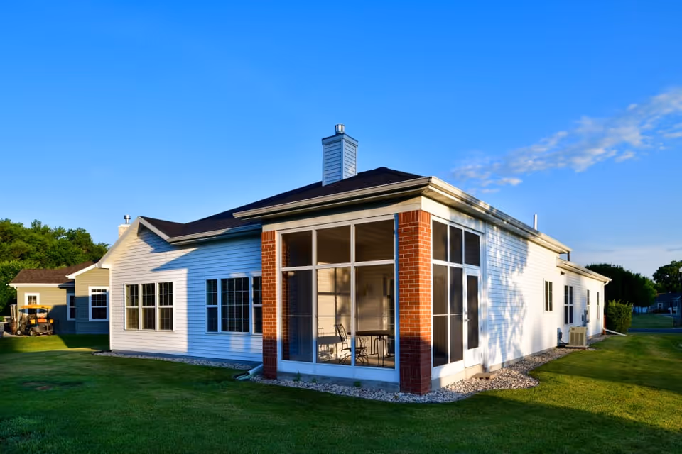 Single-story white siding building with a screened porch and brick columns on a green lawn under a blue sky.