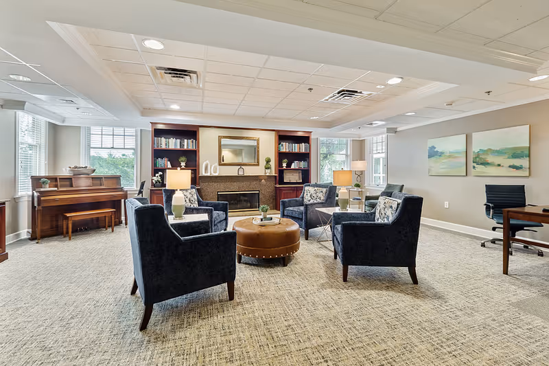 A cozy living room area with four dark blue armchairs arranged around a round brown ottoman. The room features a fireplace with a mirror above it, flanked by built-in bookshelves filled with books and decorative items. There is a wooden piano near the window on the left side, and a desk with a black office chair on the right. The walls are light-colored with two landscape paintings, and the room is well-lit with ceiling lights and table lamps.