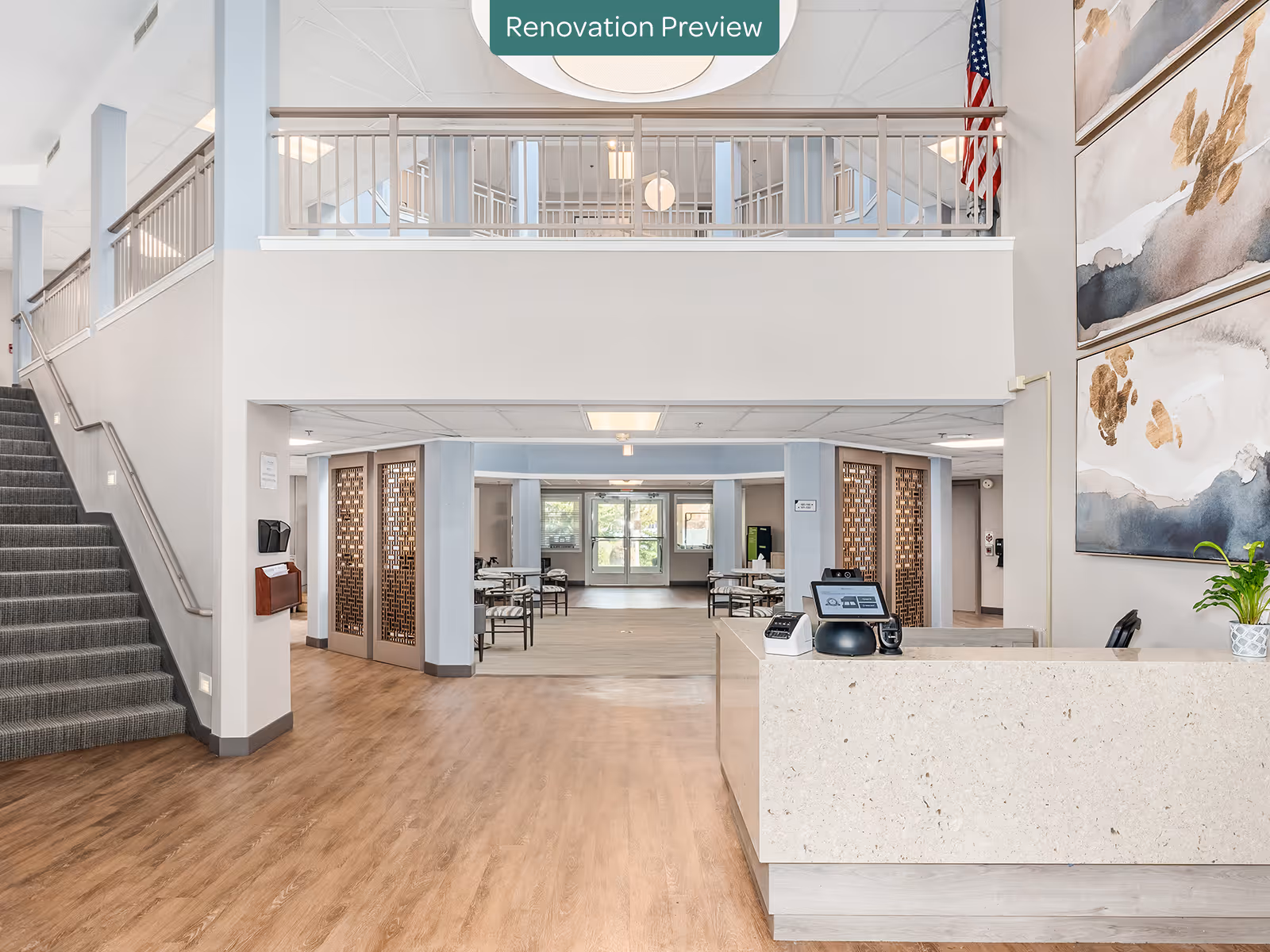 Interior view of a senior living facility lobby area with a reception desk on the right, a staircase on the left, and an open seating area with tables and chairs in the background. The space features light wood flooring, light blue and beige walls, large abstract paintings on the right wall, and an American flag near the upper railing. A sign above reads 'Renovation Preview'.
