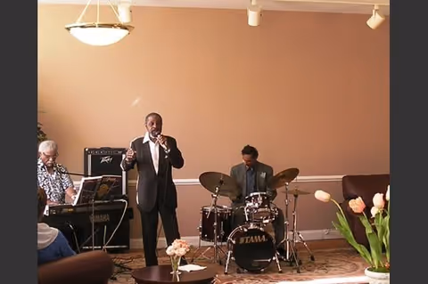 A small musical performance in a living room setting with a man singing into a microphone, a person playing a keyboard, and another playing drums. The room has beige walls, a carpeted floor, a round table with flowers, and a couch.