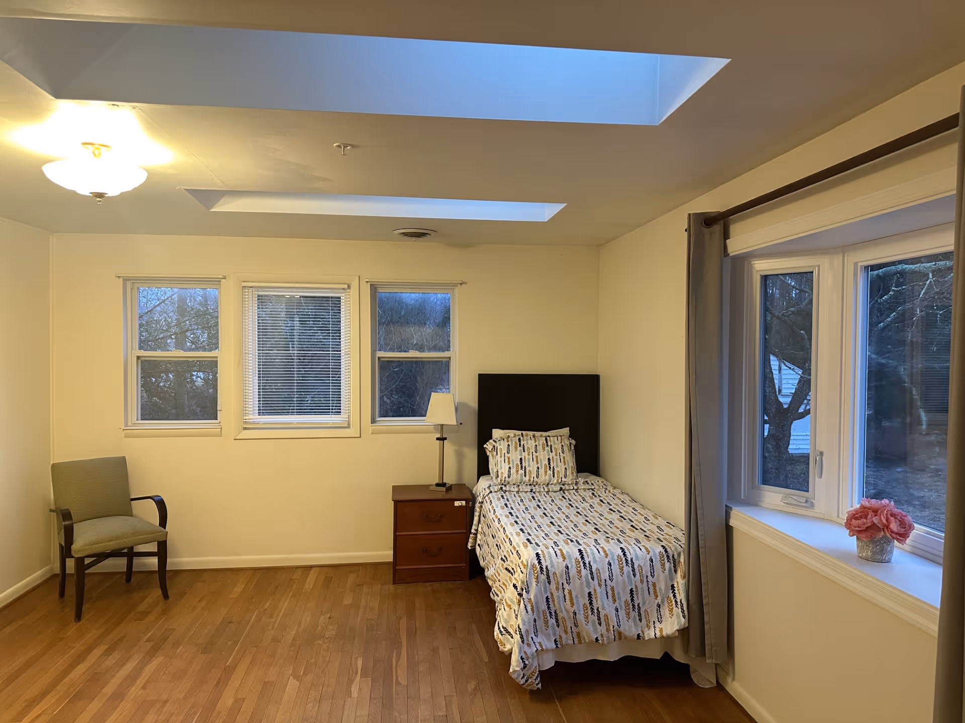 A simple bedroom with a single bed covered with a patterned bedspread, a wooden nightstand with a lamp, a green upholstered chair, three windows with blinds, and a large window with gray curtains and a small vase of pink flowers on the windowsill. The room has wooden flooring and two skylights in the ceiling.