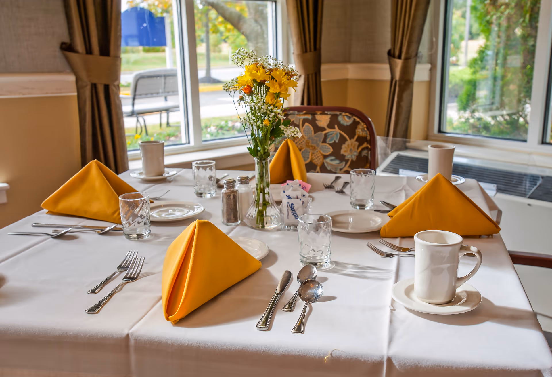 A dining table set for four with white tablecloth, yellow folded napkins, glassware, plates, silverware, and a vase with yellow and white flowers. The table is near windows with brown curtains, and outside greenery is visible.