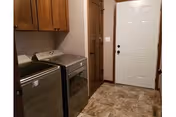 Laundry room with a stainless steel washing machine and dryer on the left side, wooden cabinets above them, a wooden door in the center, and a white exterior door on the right. The floor is covered with beige and brown patterned tiles.