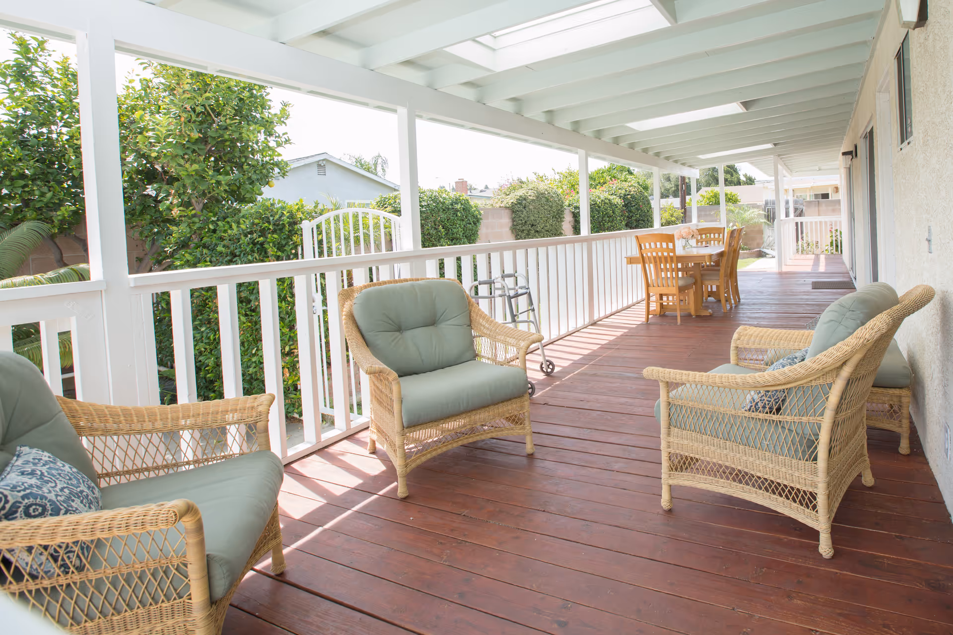 Covered outdoor porch area with wooden floor and white railing. The porch is furnished with wicker chairs with green cushions and a wooden dining table with chairs. There is a walker near the railing and greenery including bushes and trees outside the porch.