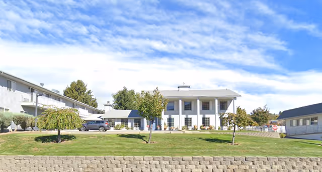 Exterior view of East Wenatchee Senior Living facility showing a large white building with columns, surrounded by green grass, small trees, and a stone retaining wall under a partly cloudy blue sky.