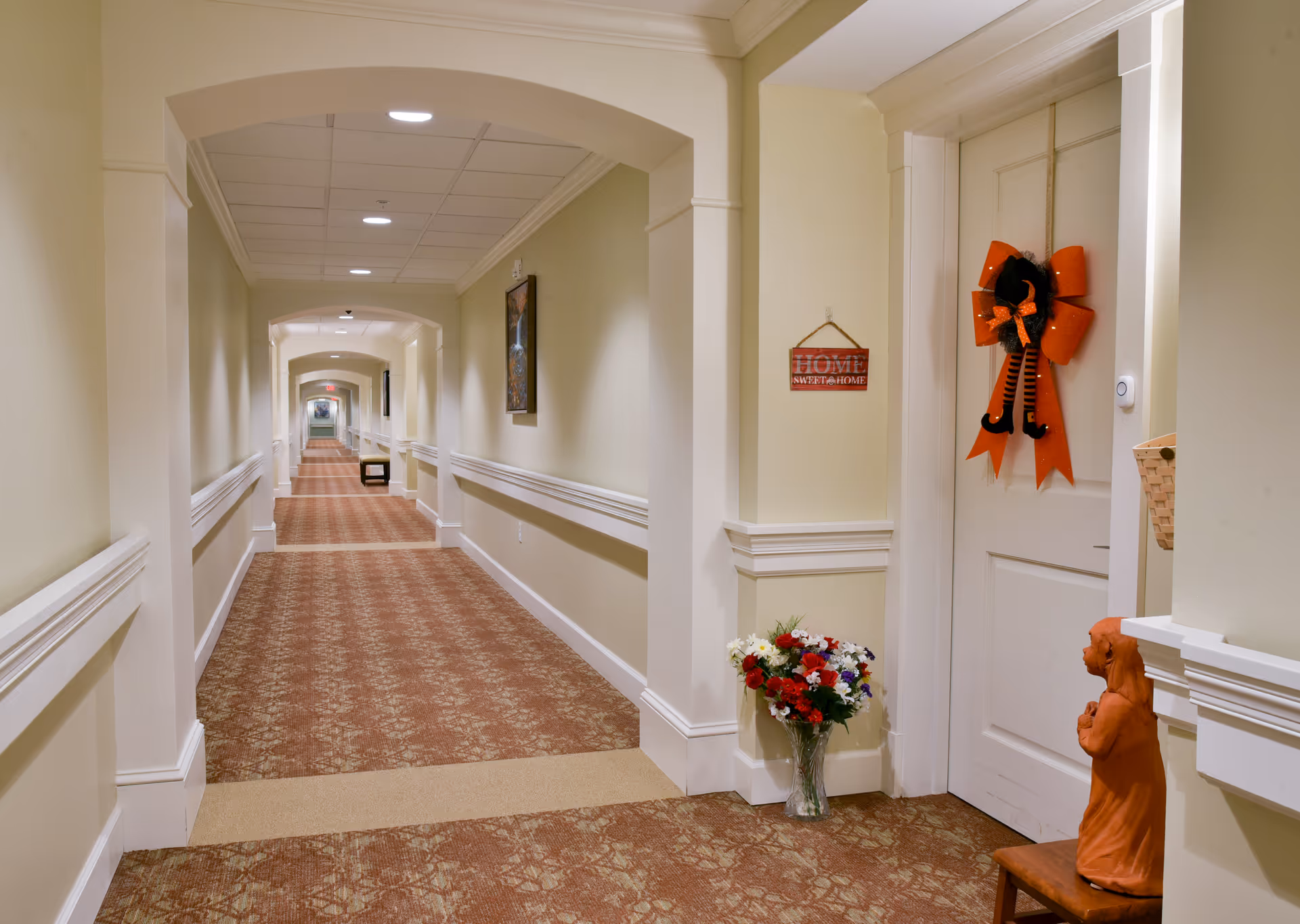 A long, well-lit hallway in a senior living facility with beige walls and patterned carpet. The hallway features white trim and handrails along the walls. On the right side, there is a white door decorated with an orange bow and a small hanging sign that reads 'HOME SWEET HOME.' Below the sign is a vase with a bouquet of colorful flowers and a small statue of a dog sitting on a wooden stool.
