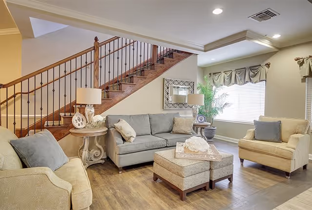 A cozy living room area featuring a gray sofa, two beige armchairs, and a matching ottoman with a decorative tray on top. The room has wooden flooring, a staircase with wooden railing, a large window with curtains, two side tables with lamps, a wall mirror, and a potted plant near the window.
