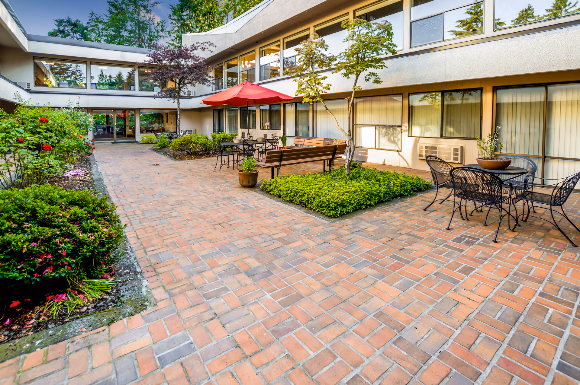 Outdoor courtyard area of Greenridge Estates Assisted Living featuring brick-paved walkways, garden beds with green shrubs and flowers, several metal tables and chairs, a wooden bench, and a red patio umbrella. The courtyard is surrounded by a two-story building with large windows.