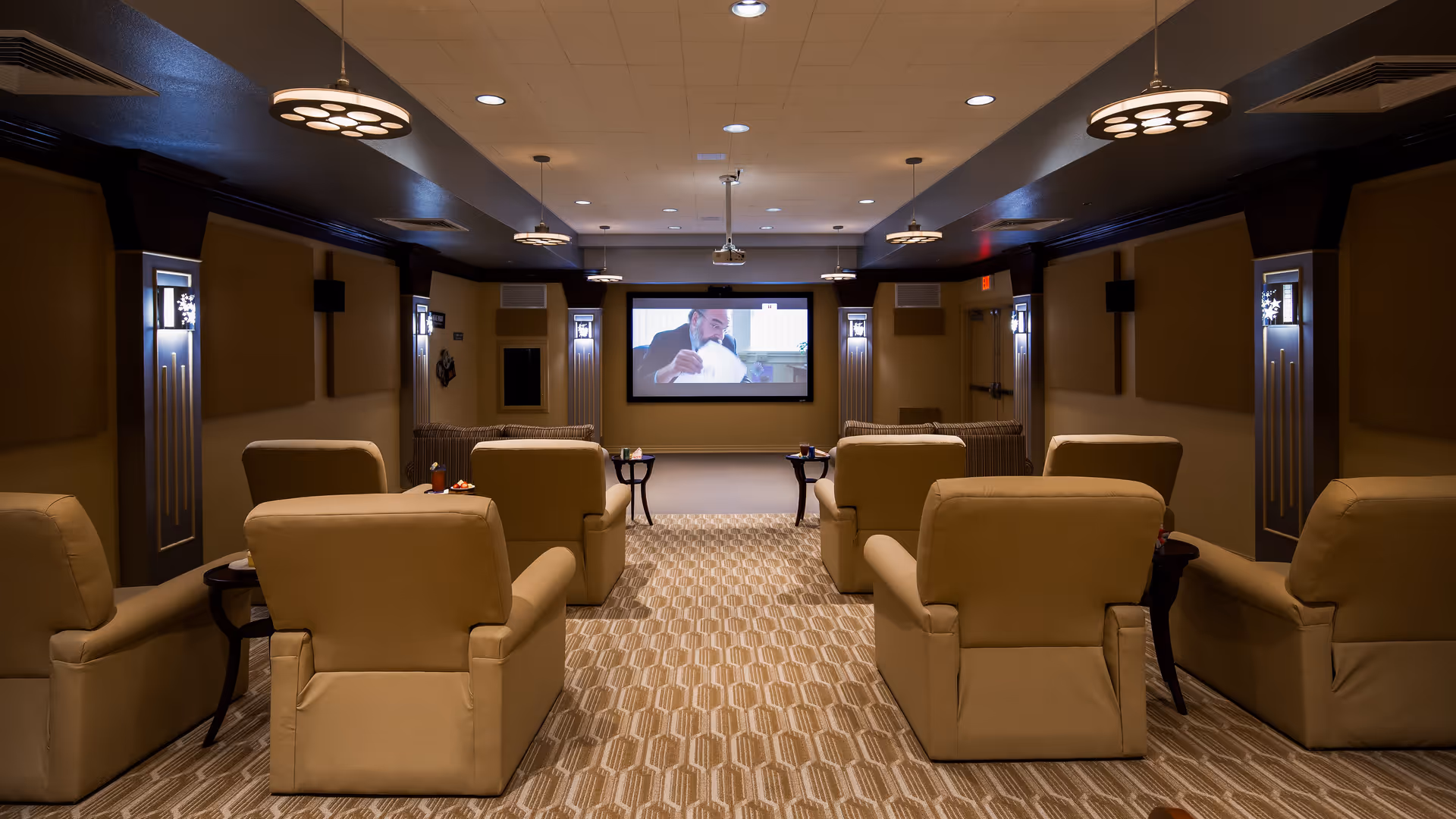 A cozy home theater room with beige recliner chairs arranged in two rows facing a large screen on the wall. The room has warm lighting with ceiling fixtures and wall sconces, and a patterned carpet covering the floor.