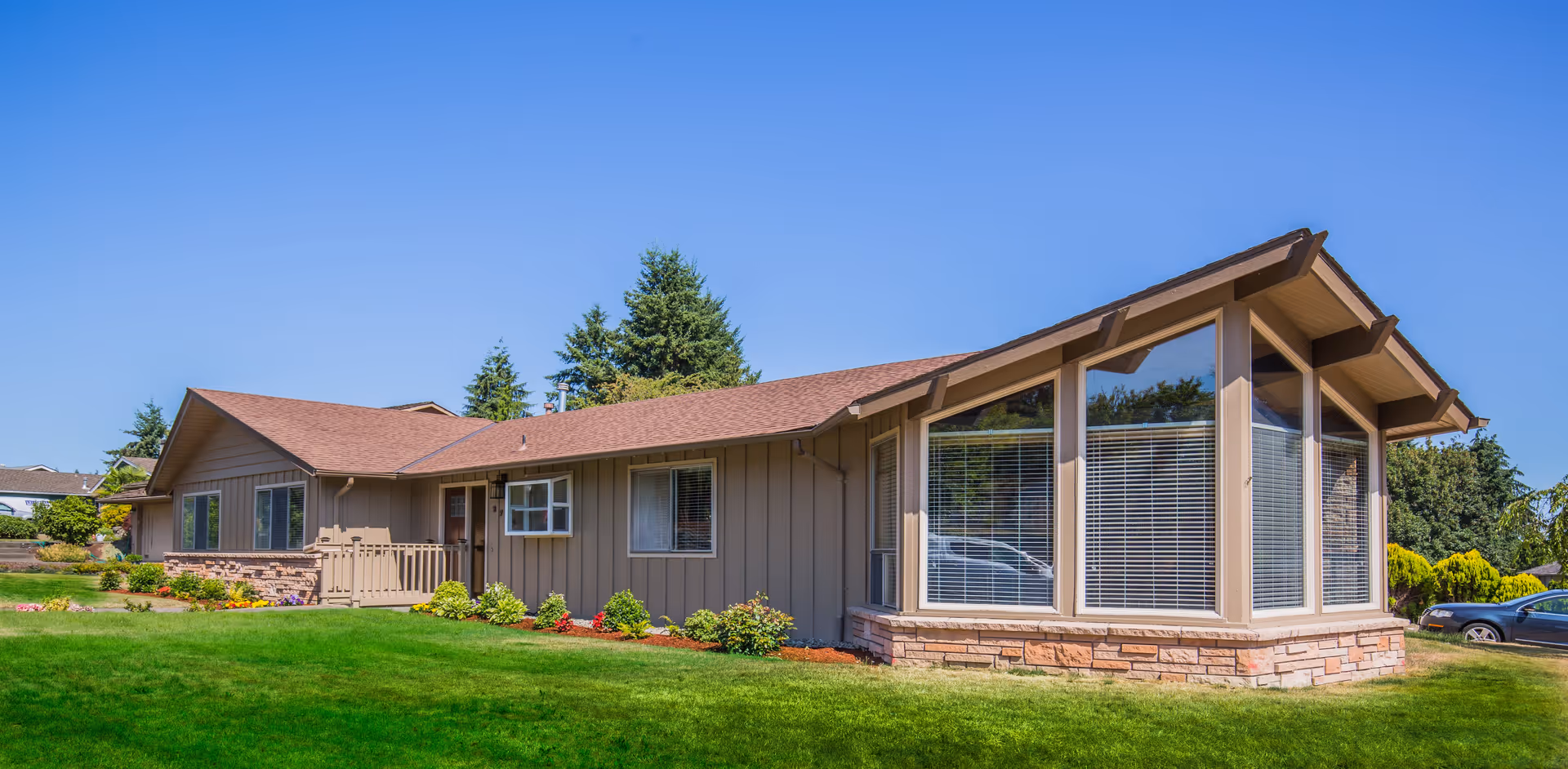 Single-story residential building with beige siding and a brown roof, featuring large windows with blinds on the right side, surrounded by a well-maintained green lawn and some shrubs under a clear blue sky.