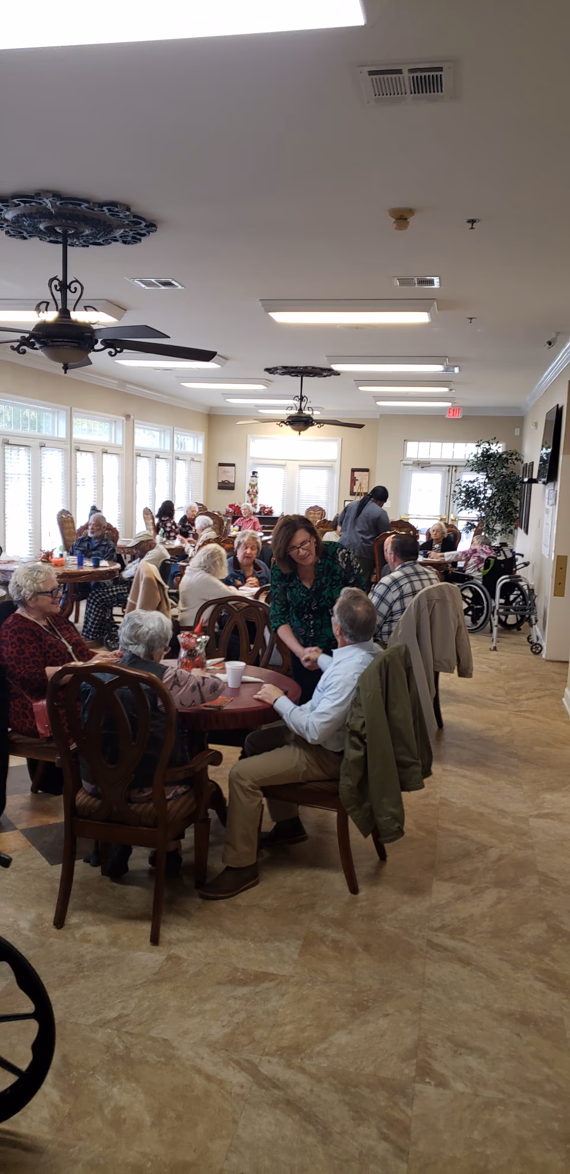 Seniors and staff gathered around tables in a bright, open communal dining/activity room.