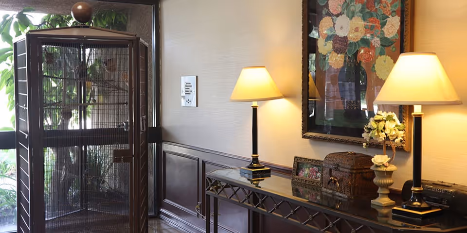 Interior view of a room with a large birdcage on the left side near a window. On the right side, there is a glass-top console table with two lit table lamps, a framed floral painting on the wall, a small decorative chest, a photo frame, and a flower arrangement in a vase.