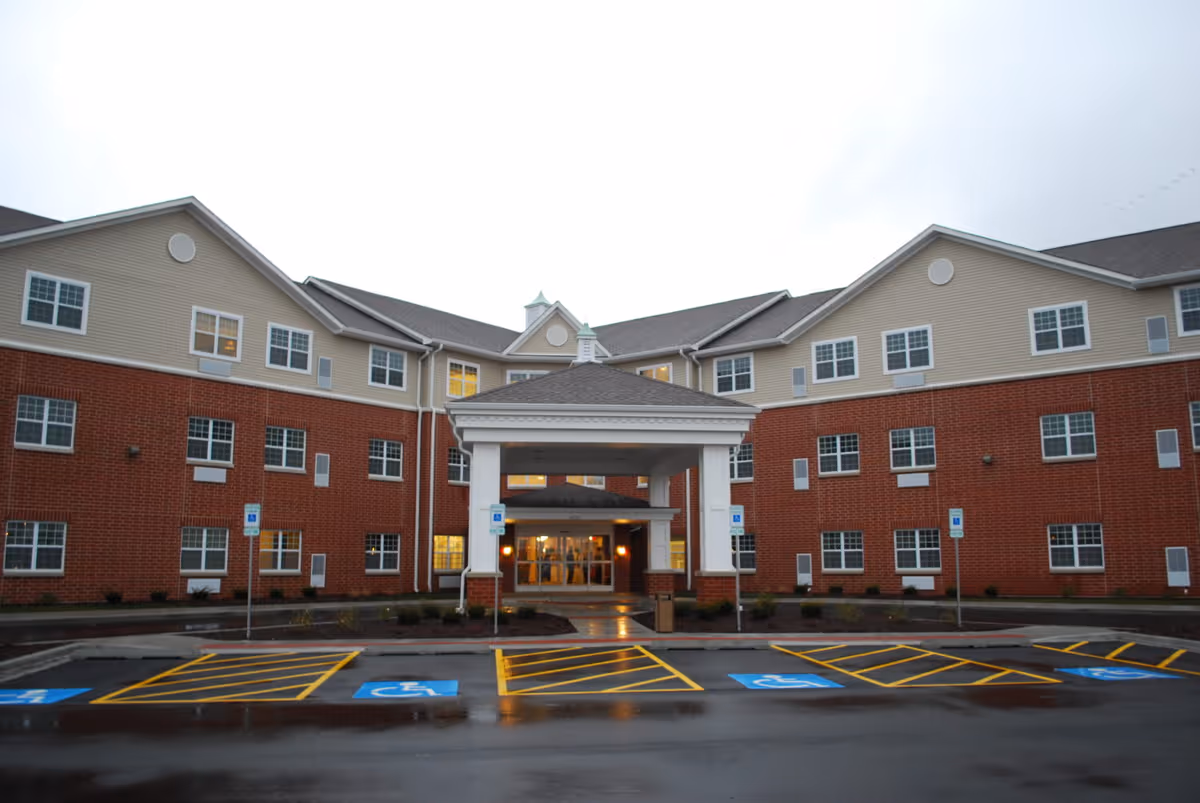 Front entrance of a three-story brick-and-siding senior living facility with a covered portico and marked handicapped parking spaces.
