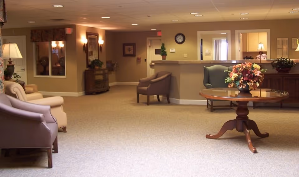 A spacious, warmly lit interior common area with beige walls and carpet. The room features several upholstered chairs arranged along the walls, a round wooden table with a floral arrangement in the center, a reception desk in the background, and decorative lamps and framed artwork on the walls.