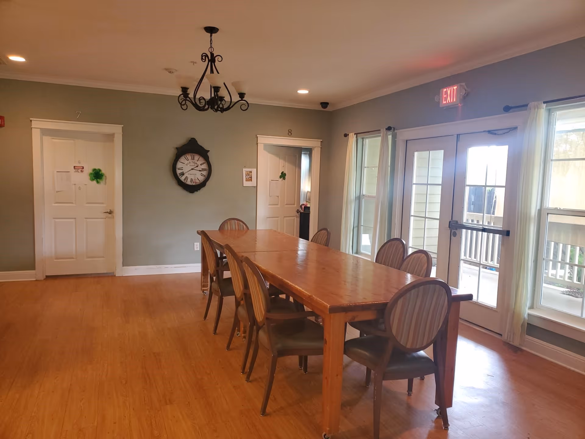 A dining room with a long wooden table surrounded by eight chairs with striped upholstery. The room has light green walls, two white doors numbered 7 and 8, a large wall clock, a chandelier, and large windows with sheer curtains letting in natural light. There is an exit door with a red exit sign above it.