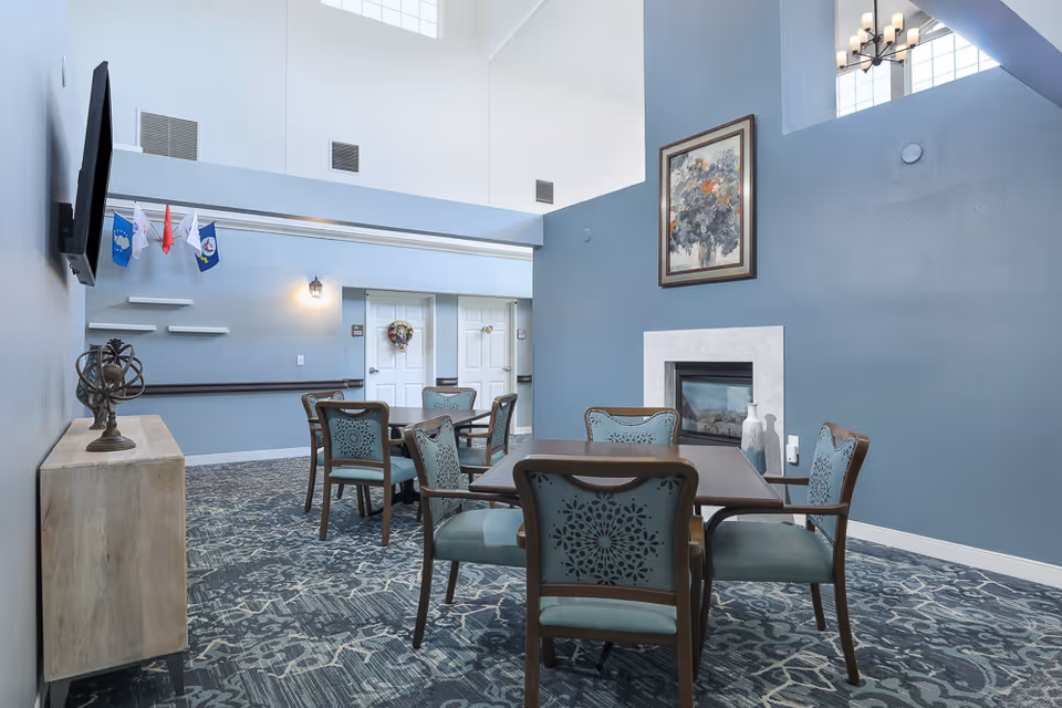A common area in an assisted living facility with blue walls and patterned carpet. There are two square tables each surrounded by four wooden chairs with blue upholstered seats and backs. A fireplace with a white mantle and a framed floral painting above it is on the right wall. On the left wall, a flat-screen TV is mounted above a wooden console table with a decorative sculpture. Small flags hang on the wall near two white doors in the background.