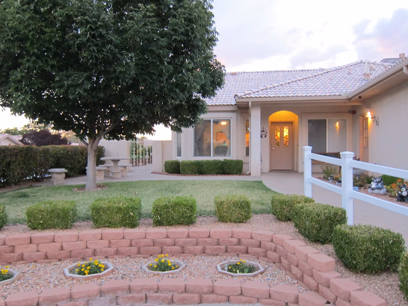 Front yard of a single-story building with a tiled roof, featuring a well-maintained garden with bushes, a tree, and flower beds bordered by red bricks. A white fence runs along the right side of the walkway leading to the front door, which is illuminated by a porch light.
