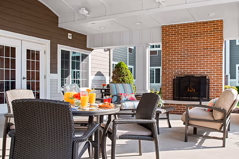 Outdoor covered patio area with a round table set for breakfast including orange juice and pastries, surrounded by wicker chairs. There are cushioned lounge chairs near a brick fireplace, and the area is part of a residential building with white columns and brown siding.
