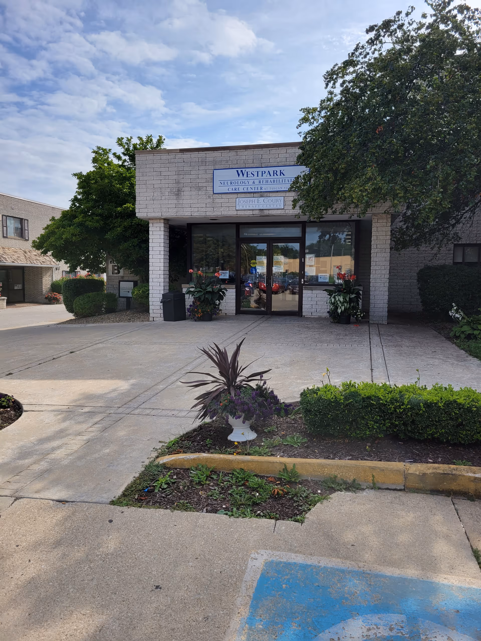 Front entrance of Westpark Neurology & Rehabilitation Care Center with a concrete driveway, potted plants, and greenery on either side. The building is made of light-colored bricks with glass doors and a sign above the entrance. A tree partially shades the right side of the entrance.