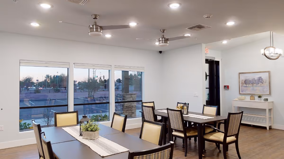 Bright communal dining room with dark wood tables, cushioned chairs, large windows, ceiling fans, and simple wall decor.