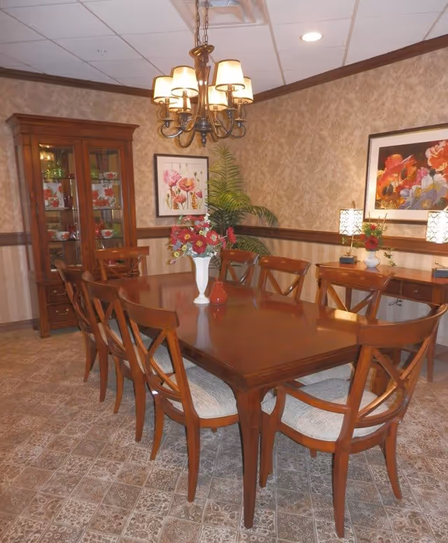 A formal dining room with a long wooden table surrounded by eight wooden chairs with cushioned seats. A white vase with red and pink flowers is placed in the center of the table. The room features patterned wallpaper, a chandelier with six lampshades hanging above the table, a wooden cabinet with glass doors displaying glassware, framed floral artwork on the walls, two table lamps on a wooden sideboard, and a potted plant in the corner.