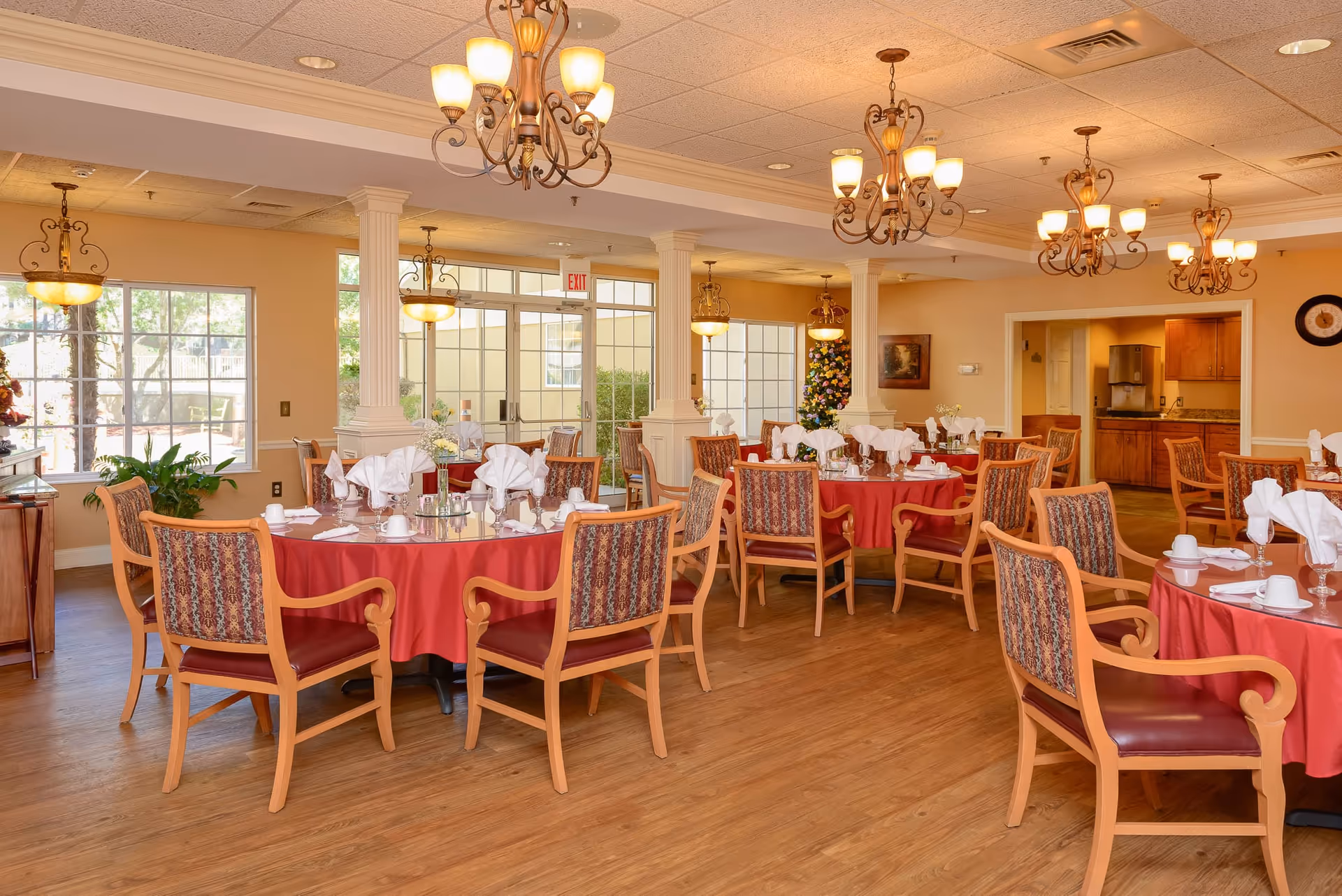 A dining room in a senior living facility with round tables covered in red tablecloths, each set with white napkins, cups, and glassware. The room has wooden chairs with patterned upholstery, chandeliers hanging from the ceiling, large windows letting in natural light, and a door leading outside. The walls are painted a warm beige color, and there are decorative columns and plants near the windows.