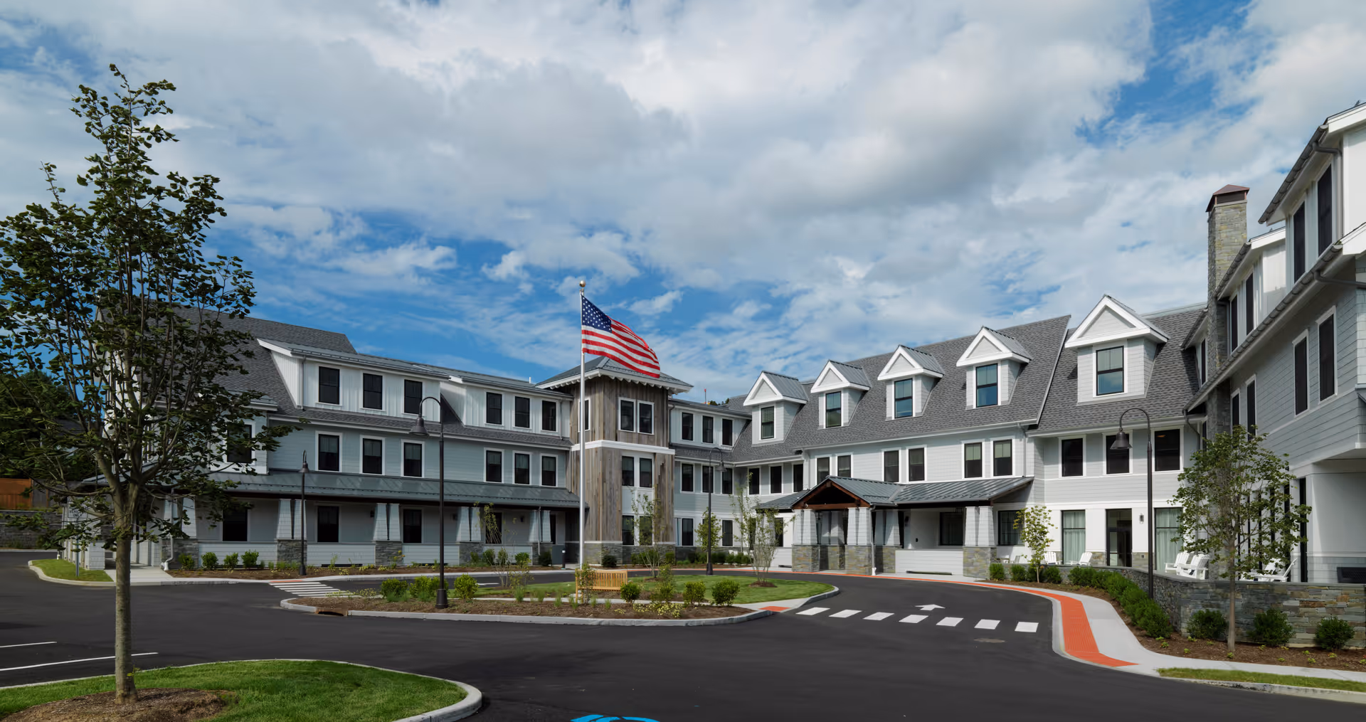 Exterior view of a large, modern senior living facility with white siding and multiple windows. The building has a central tower with an American flag flying on a flagpole in front. There is a circular driveway with landscaped greenery and young trees under a partly cloudy sky.