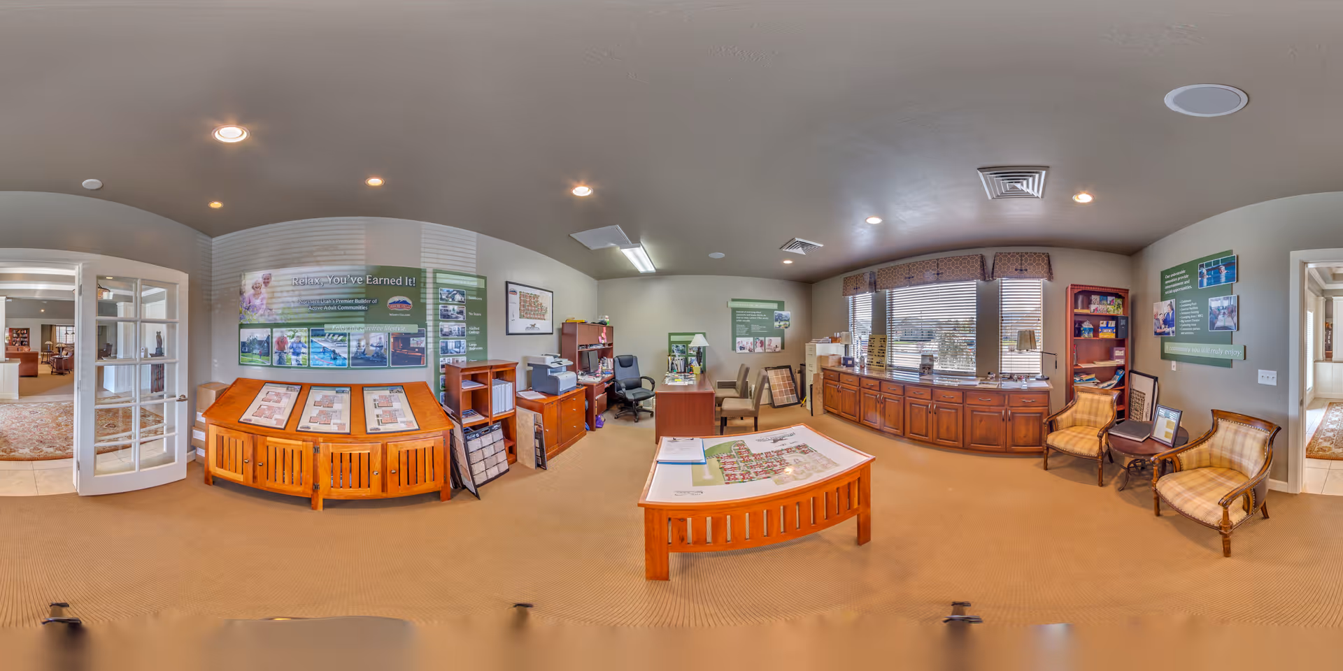 A well-lit office or reception area with beige walls and carpet. The room features wooden furniture including cabinets, a desk, and display tables with brochures and floor plans. There are two upholstered chairs near a window with blinds, and informational posters on the walls. A glass door leads to another room with more seating visible.