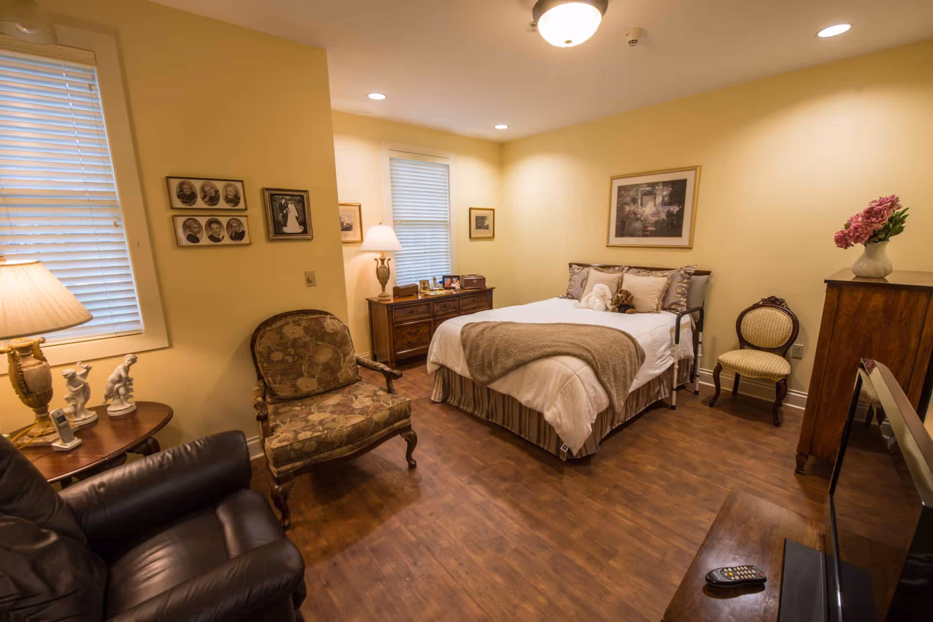 Furnished bedroom with a bed, bedside tables and lamps, seating area with armchairs, dresser, and warm wood flooring.