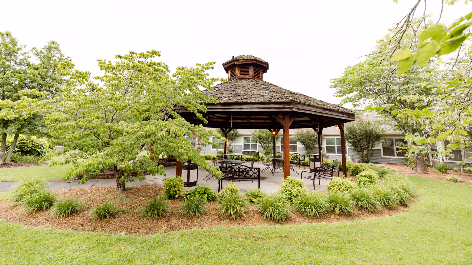 A wooden gazebo with a shingled roof surrounded by green shrubs and trees in a well-maintained garden area, with benches and chairs placed inside the gazebo. A building with multiple windows is visible in the background.