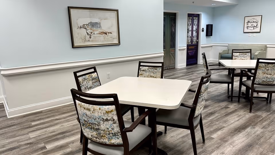 A clean and modern dining area with two square white tables surrounded by chairs with patterned backs and gray seats. The room has light blue walls with white trim, wood-look flooring, a framed artwork of a dog on one wall, and a framed sign that says 'forever grateful' on another wall. There is also a small gray couch in the background near a doorway with decorative glass panels.