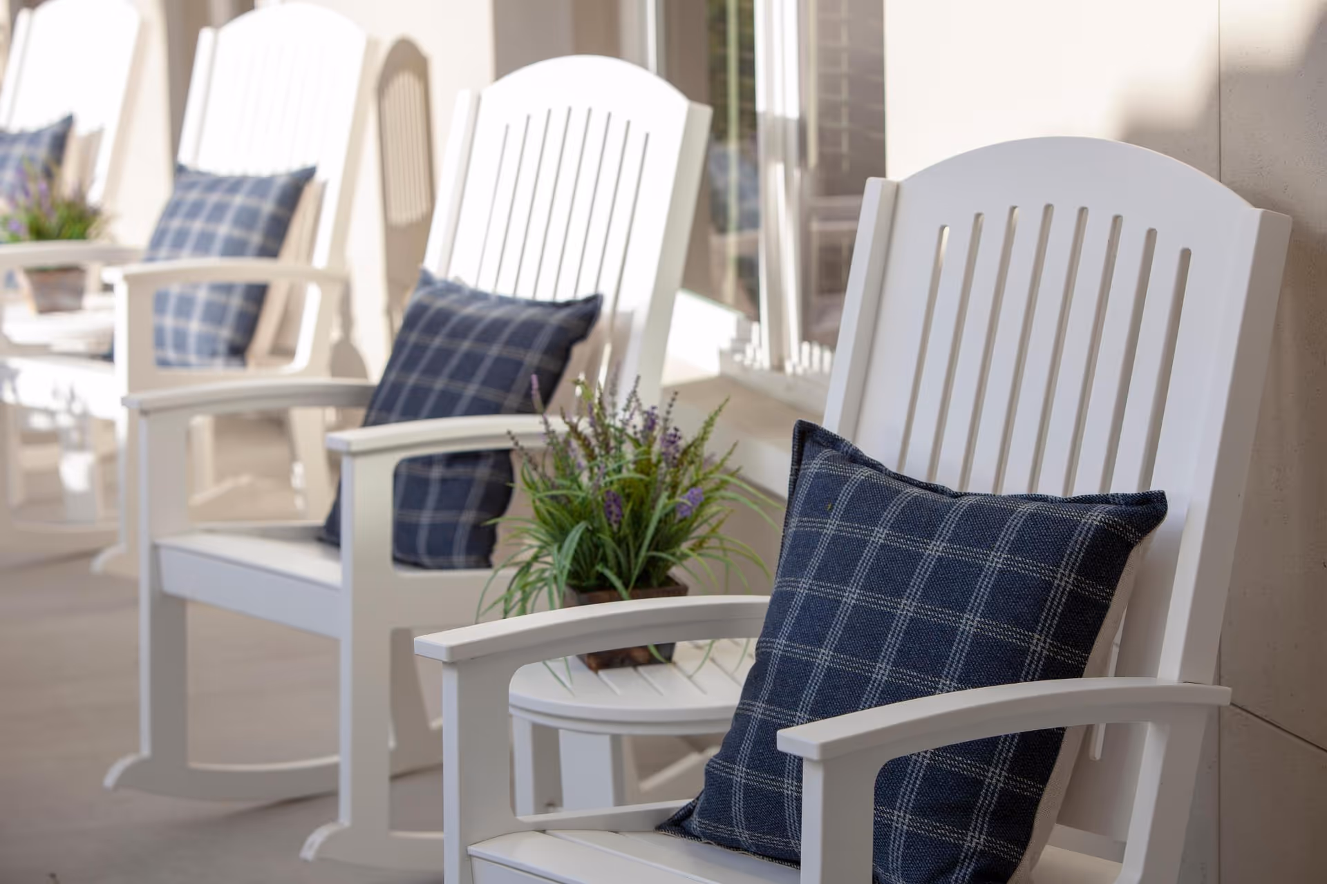 A row of white wooden rocking chairs with blue plaid cushions on a covered porch. A small round table with a potted plant is placed between the chairs. The setting is bright and inviting with sunlight streaming in.