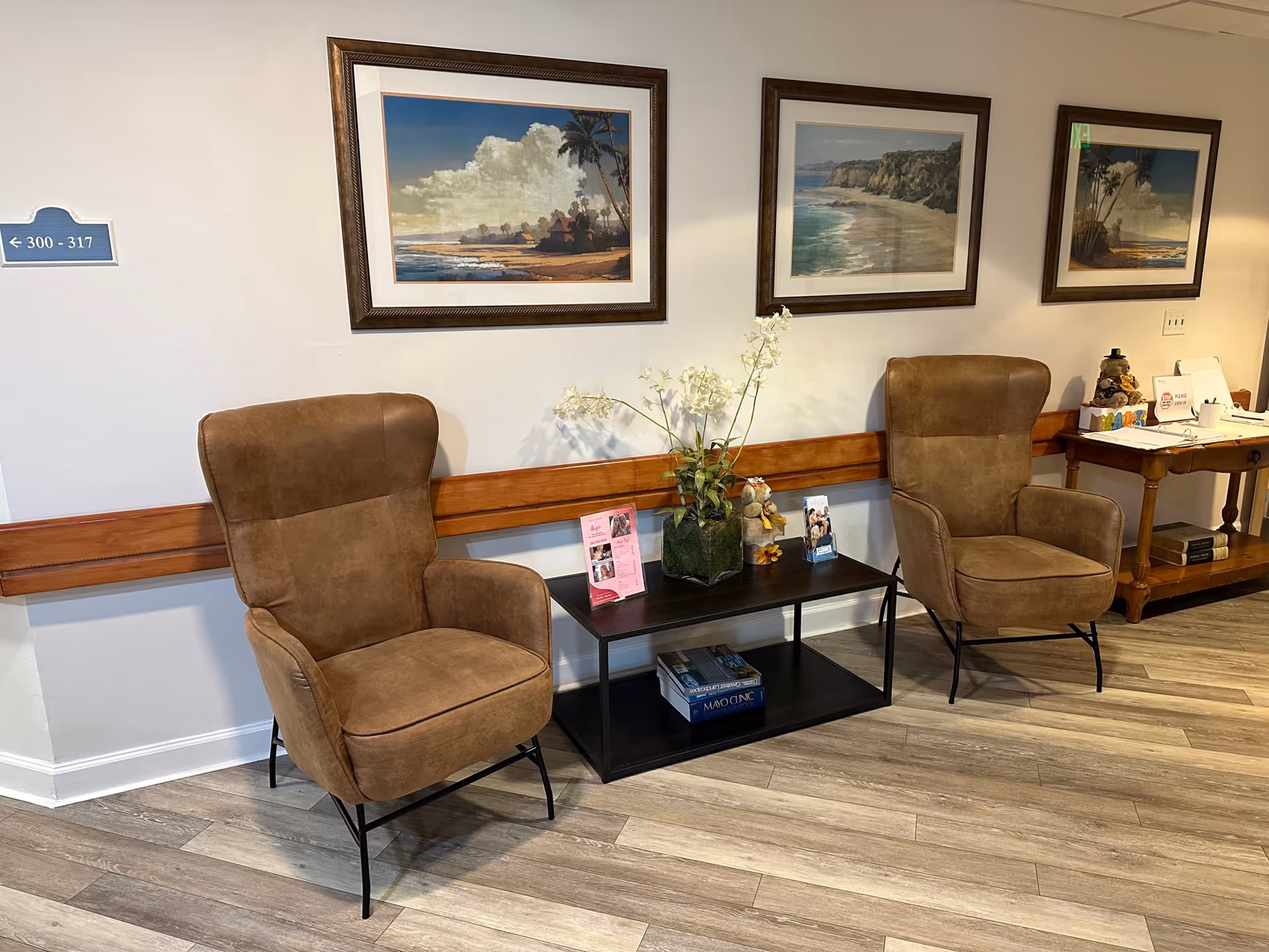Two brown armchairs flank a small black table with a plant and magazines beneath framed coastal paintings on a facility interior wall.