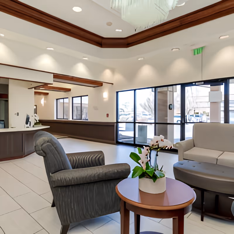 A bright and spacious senior living facility lobby with a gray armchair and a beige sofa arranged around a small round wooden table with a potted plant. Large windows and glass doors allow natural light to fill the room, and a reception desk is visible in the background.