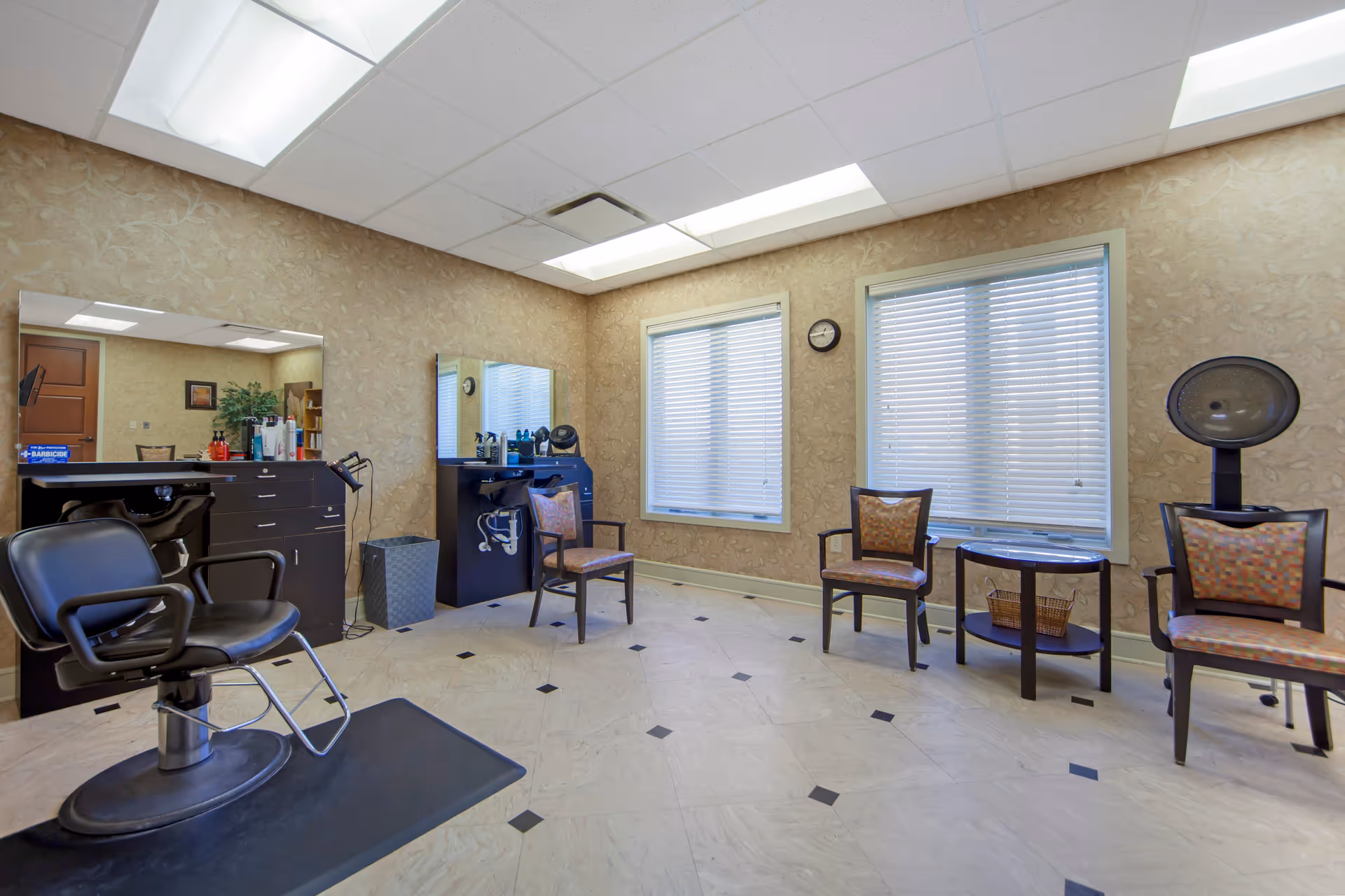 Interior view of a hair salon area in an assisted living facility featuring a black salon chair on a mat, two large mirrors above black counters with hair care products, three patterned chairs, a small round table with a basket underneath, two windows with blinds, and a hair dryer station. The room has beige patterned wallpaper and a tiled floor with black diamond accents.