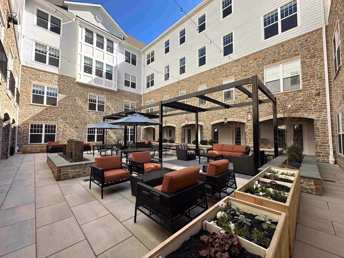 Sunlit enclosed courtyard with patio seating, pergolas and planters surrounded by a multi-story stone and siding building.