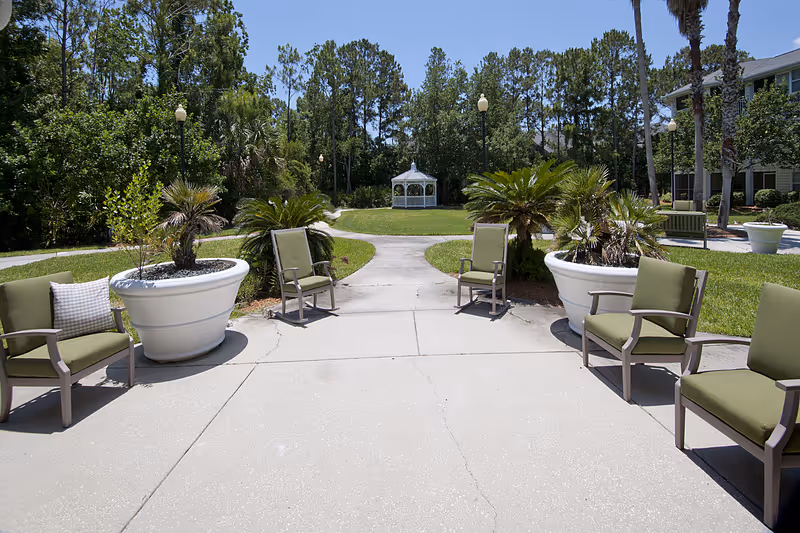 Outdoor patio area with green cushioned chairs arranged around large white planters containing small palm plants. A concrete pathway leads to a white gazebo in the background surrounded by trees and greenery under a clear blue sky.